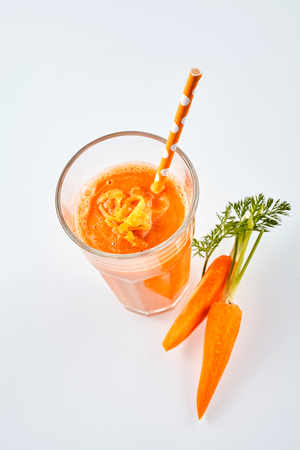 Overhead view of one large glass of orange root vegetable beverage with a straw next to fresh carrots on a white backgroundの写真素材