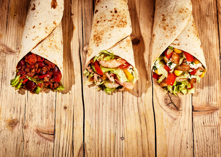 Front view of three meat, bean and vegetable stuffed burritos in baked wheat tortillas on old wooden table tableの写真素材