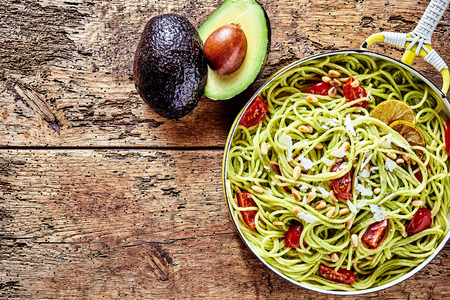 Exotic Italian spaghetti pasta with avocado pear, pine nuts, tomato and parmesan cheese served in a pan on a rustic wooden table with copy space, viewed from aboveの写真素材