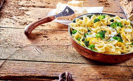 Serving of farfalle, bow-tie or butterfly pasta with fresh green broccoli and grated parmesan cheese on a rustic wooden counter with a block of cheese and cutter in the background, copy spaceの写真素材