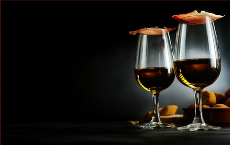 Spanish tapas and sherry aperitif with slices of gourmet ham balancing on top of the glasses with a bowl of bacalao croquettes over a dark background with copy spaceの写真素材