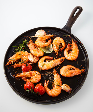 Delicious grilled spicy prawns seasoned with rosemary, fresh garlic, lime and cherry tomatoes viewed from overhead in an old rustic frying pan on whiteの写真素材