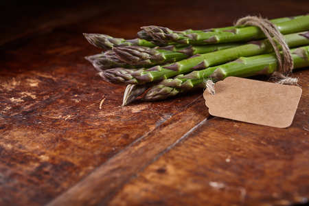 Selective focus close up view of green asparagus tips wrapped at stem with string and blank label with copy space over rough wooden tableの写真素材