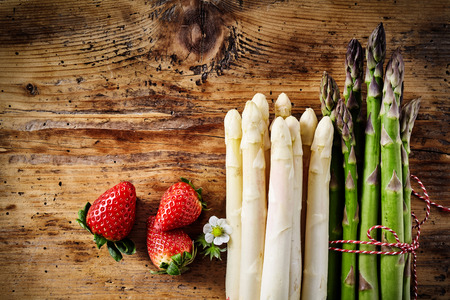 Three ripe red strawberries with stemmed floret along side of bundled white and green asparagus on wooden tableの写真素材