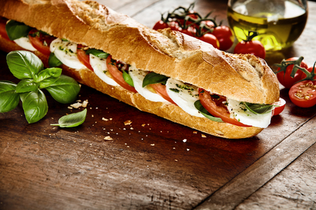 Preparing a fresh crusty baguette with Caprese filling in the kitchen with fresh basil leaves, cherry tomatoes, olive oil and mozzarella cheeseの写真素材