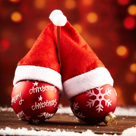 Cozy pair of colorful red Christmas baubles with Santa hats and decorated motifs on a wooden table with snow against a background of warm sparkling party light bokehの写真素材