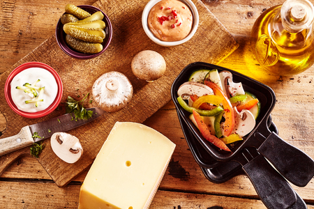 Overhead view of the fresh raw ingredients for a delicious raclette cheese recipe with diced fresh vegeables, olive oil, mushrooms, herbs, gherkins and a large wedge of Swiss racletteの写真素材