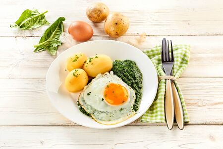 Fried egg, creamy pureed spinach and baby potatoes garnished with chopped parsley on a rustic white wooden table with fresh ingredients behind and a napkin with utensilsの写真素材