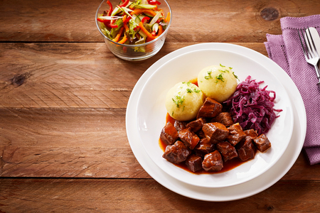 Traditional German cuisine with tasty rich beef goulash and boiled dumplings served with shredded purple cabbage and a fresh salad, rustic wooden boards and copy spaceの写真素材