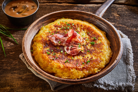 Delicious golden big pan potato fritter, an Austrian speciality recipe garnished with fresh herbs and a rasher of ham or bacon, high angle close up viewの写真素材