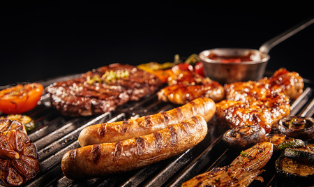 Barbecuing an assortment of meat with pork sausages, beef steak, chicken wings onions, tomatoes and...