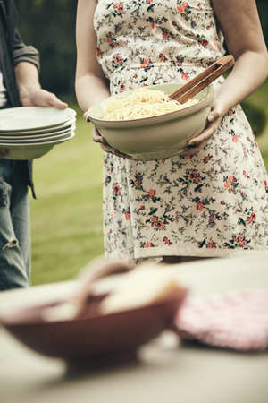 Young woman bringing a bowl of spaghetti to the table outdoors while her boyfriend carries a stack of plates in a catering conceptの写真素材