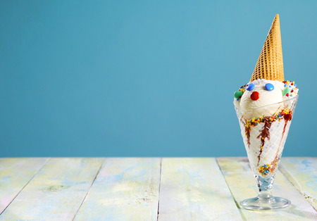 Fun ice cream sundae for a kids party with a cute little clowns face decorated with sugar coated candy and a cone for a hat on a rustic table with copy space over blue