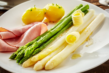 Serving of fresh green and canned white asparagus spears garnished with melting butter and served with sliced gammon and baby potatoesの写真素材