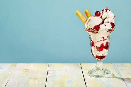 Close Up Still Life of Sundae Made with Scoops of Vanilla Ice Cream, Cherries, Biscuits and Whipped Cream Served in Glass Dish on Painted Wood Table with Blue Background and Copy Spaceの写真素材
