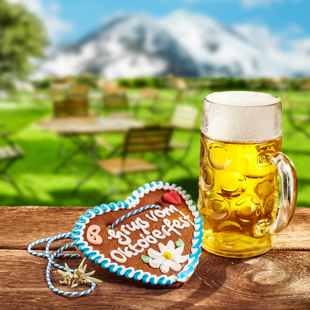 Oktoberfest concept with a ginger heart with decorative icing and a glass tankard of beer or lager outdoors on a rustic tavern table in Bavaria with snowy mountainsの写真素材