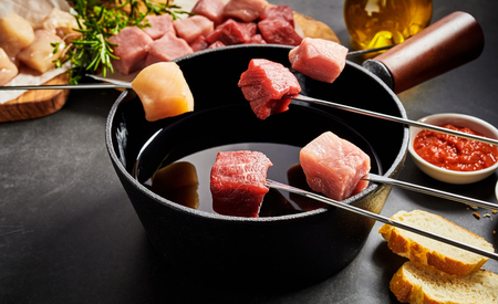 Assorted fresh lean meat ready for dipping in a fondue pot displayed on forks above the oil with ingredients, bread and dips to the sidesの写真素材