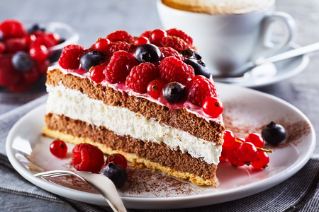 Slice of gourmet fresh berry cake on a plate with layered cream topped with fresh autumn raspberries, blueberries and redcurrants in a close up view on a plate