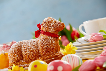 Gourmet speciality lamb shaped Easter cake with red bow on a festive table with colorful eggs, spring daffodils and stacked plates for servingの写真素材