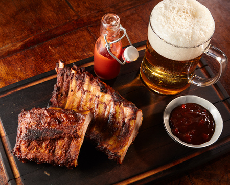 Grilled or barbecued spicy ribs with a cold beer with frothy head served on a tray with chutney and tomato ketchup for a tasty finger snack pub lunchの写真素材