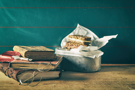 Vintage still life of a school lunch break with a wholegrain sandwich wrapped in paper on an old tin container alongside worn leather books and spectaclesの写真素材
