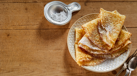 Banner or Panorama Plate of golden fried pancake wraps dusted with castor sugar viewed high angle on a wooden table with a strainer or sugar spoon and copy spaceの写真素材