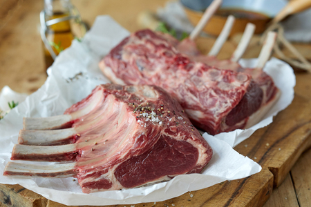 Close up on two trimmed seasoned racks of lamb standing ready for cooking on oven paper on a wooden chopping board with bone-in chops