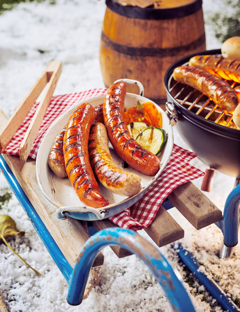 Grilled pork sausages at a winter barbecue grilling over the hot coals and heaped in a metal dish ready for serving on an old simple toboggan in fresh white snowの写真素材