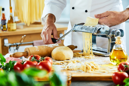 Adult male chef making noodles on top of cutting board next to other vegetables with large kitchen in backgroundの写真素材