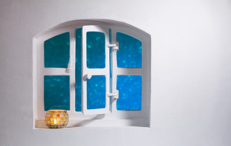White wall with open arched window with blue glow outside and candle burning in bowl on the sill. Copy spaceの写真素材