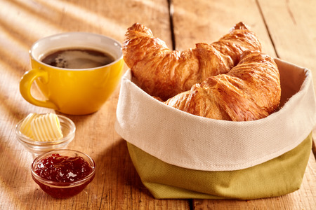 Freshly baked flaky croissants served in fabric bag with butter curls, jam and cup of coffee on rustic wooden table