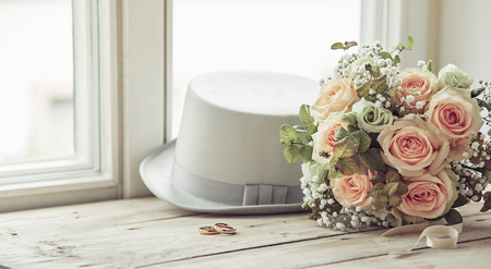 Marriage day composition of groom's white hat, wedding rings and bridal bouquet of pink roses, sitting on wooden window sillの写真素材