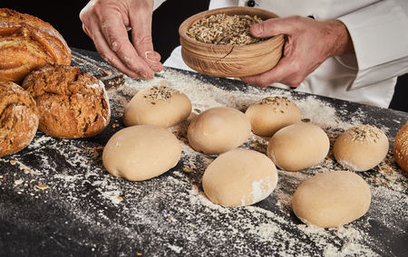 Baker sprinkling crushed grain and seeds on buns dough already formed into buns or rolls on a...