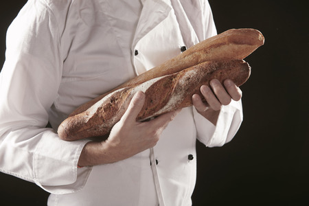 Baker in white uniform holding or carrying two baguettes of freshly baked rye bread, viewed in close-up against blackの写真素材