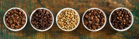 Panorama banner with assorted fresh roasted and raw coffee beans in individual bowls on rustic wood...