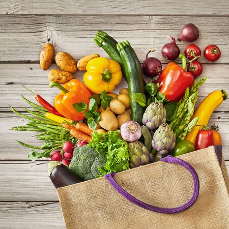 Grocery bag with purple handle that is spilling vegetables while on top of rustic wooden bench backgroundの写真素材