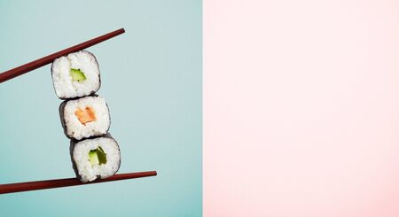 Chopsticks holding three seaweed wraps with rice and avocado over blue half of two tone background.の写真素材