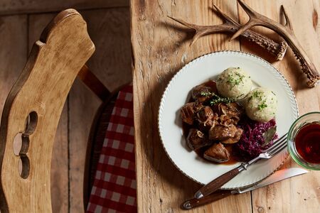 Serving of wild venison goulash with dumplings and red cabbage garnished with chopped fresh herbs on a rustic table set with deer antlers and wineの写真素材