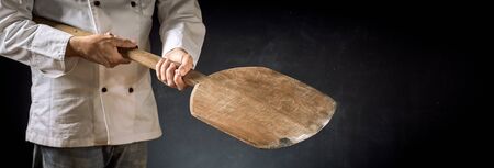 Cook or chef in a pizzeria holding an empty wooden paddle in a close up on his hands over a dark...