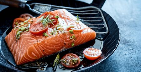 Fresh filleted salmon steak topped with chopped herbs and tomato on a small skillet with oil and spatula ready to be sauteedの写真素材
