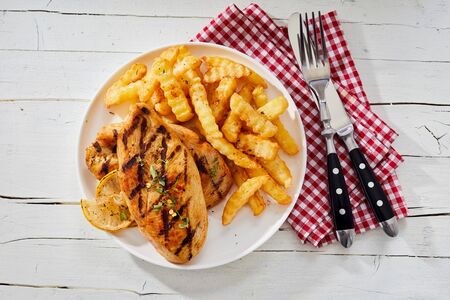Barbecued healthy skinless chicken breast seasoned with herbs and served with crinkle cut potato chips viewed from overhead with utensils and napkin on rustic white woodの写真素材