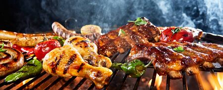 Assorted meat grilling over a BBQ outdoors in a low angle view of chicken legs, pork ribs, sausages and vegetables with rising smoke and copy space in a panorama bannerの写真素材