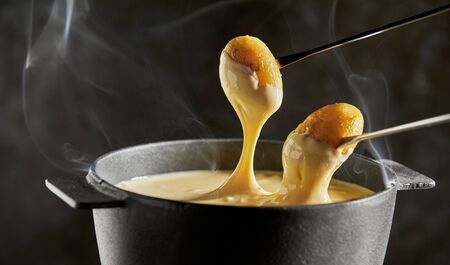 Dipping grilled baby potatoes into cheese fondue in a cast iron pot over a dark background showing the rising steam in close upの写真素材