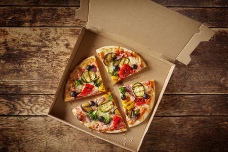 Sliced Italian pizza with fresh vegetables and salami in a cardboard fast food box for delivery on a rustic wooden table in a top down viewの写真素材