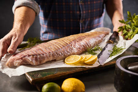 Crop unrecognizable male preparing pollock fish fillet with aromatic dill and lemon slices on kitchen tableの写真素材