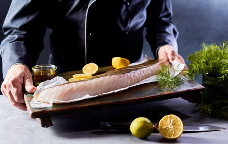 Crop unrecognizable male chef in uniform showing wooden board with uncooked organic fish fillet with lemon slices and olive oil in kitchenの写真素材