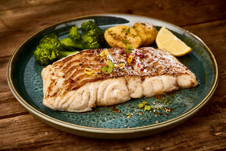 Delicious grilled pollack fish steak with crispy skin served on dark plate with steamed broccoli and...