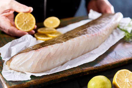 Crop unrecognizable male cook squeezing lemon juice on uncooked fresh cod fish fillet placed on wooden boardの写真素材