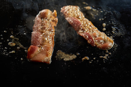From above of appetizing bacon strips preparing in hot oil of black surface in kitchenの写真素材