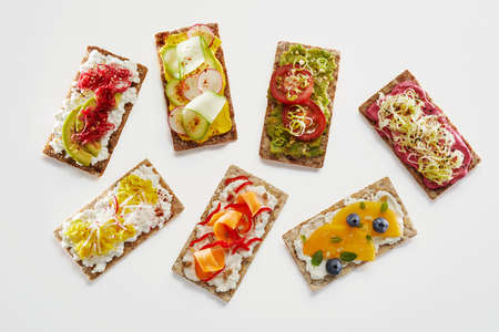 From above of assorted crispbread toasts with fresh fruits and vegetables served on white background for vegetarian snackの写真素材
